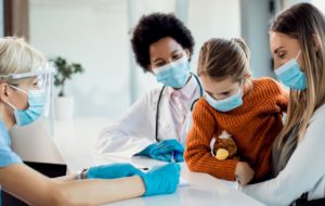 Little girl and her mother talking to a nurse at reception desk in the hospital during COVID-19 pandemic.